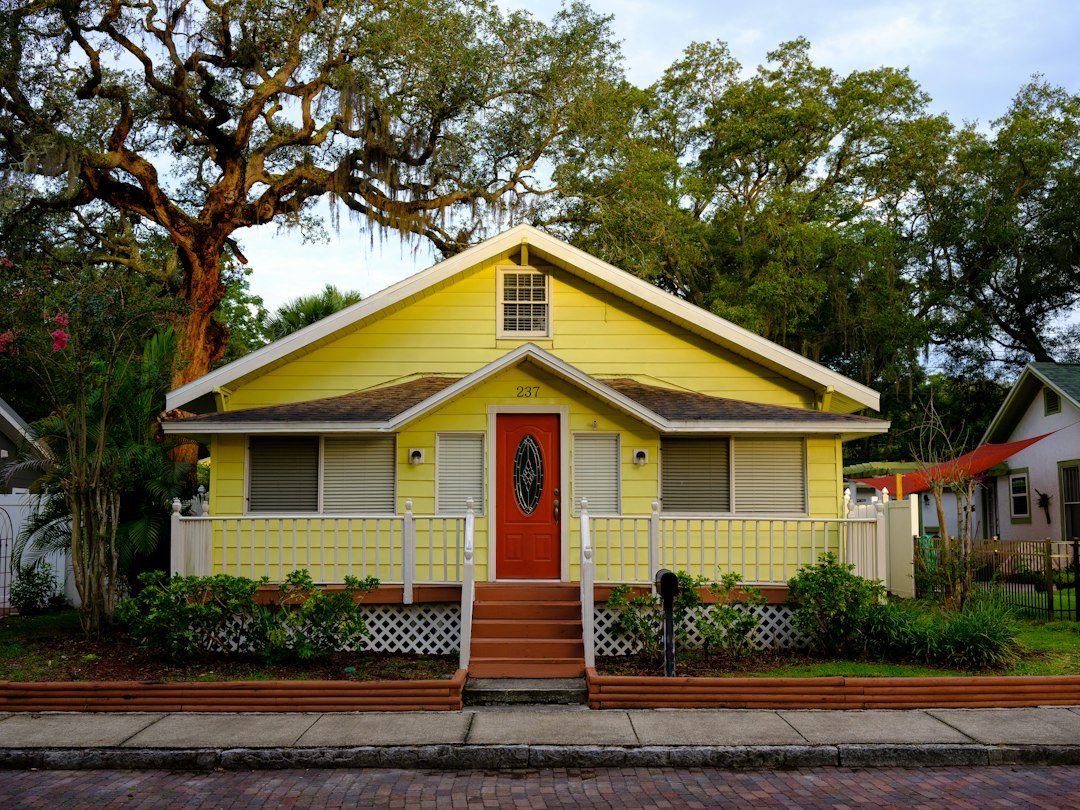 Small yellow house with a red front door, white porch railings, and trees