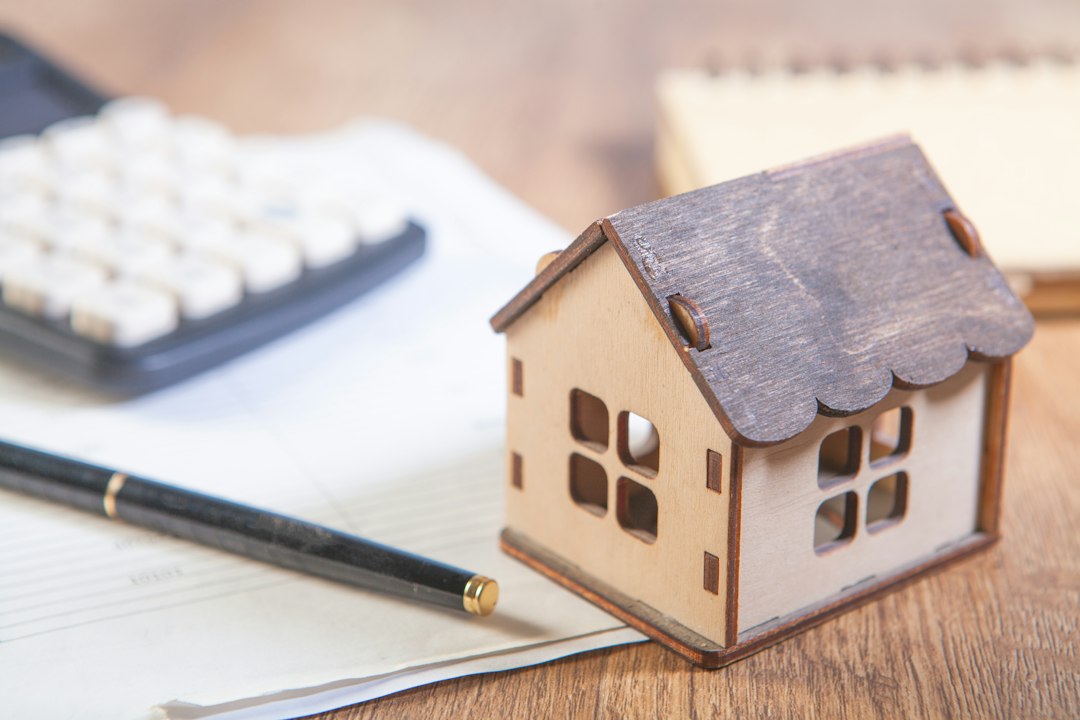 A small wooden house model beside a pencil, calculator, and paperwork.
