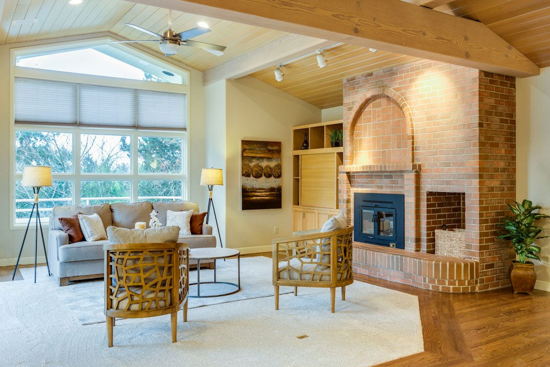 Bright family room with a brick fireplace, wood beams, wicker chairs, and tall windows