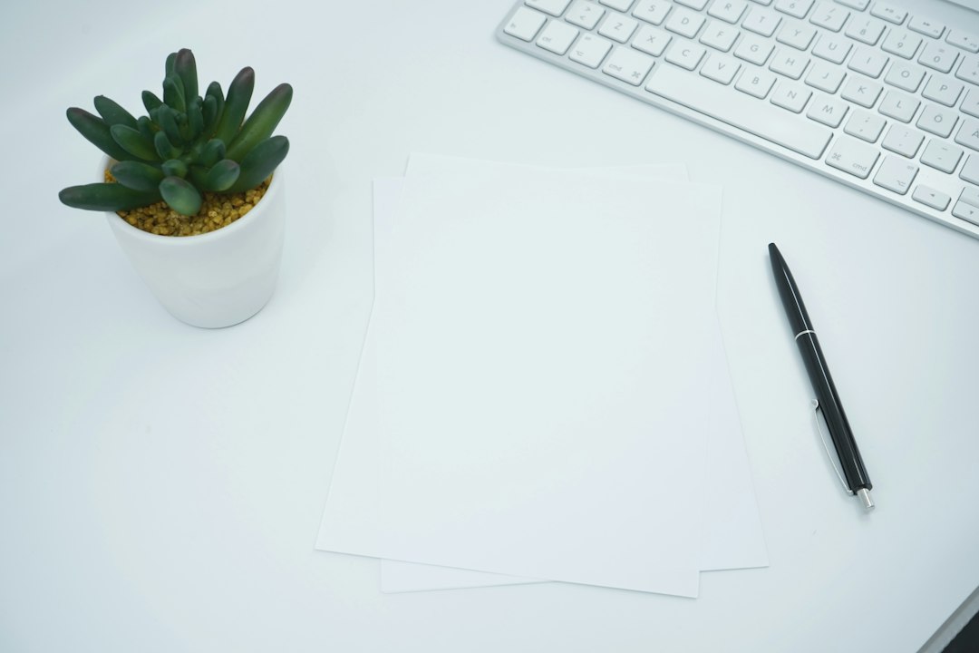 Blank papers and a black pen on a desk beside a keyboard and a small plant.
